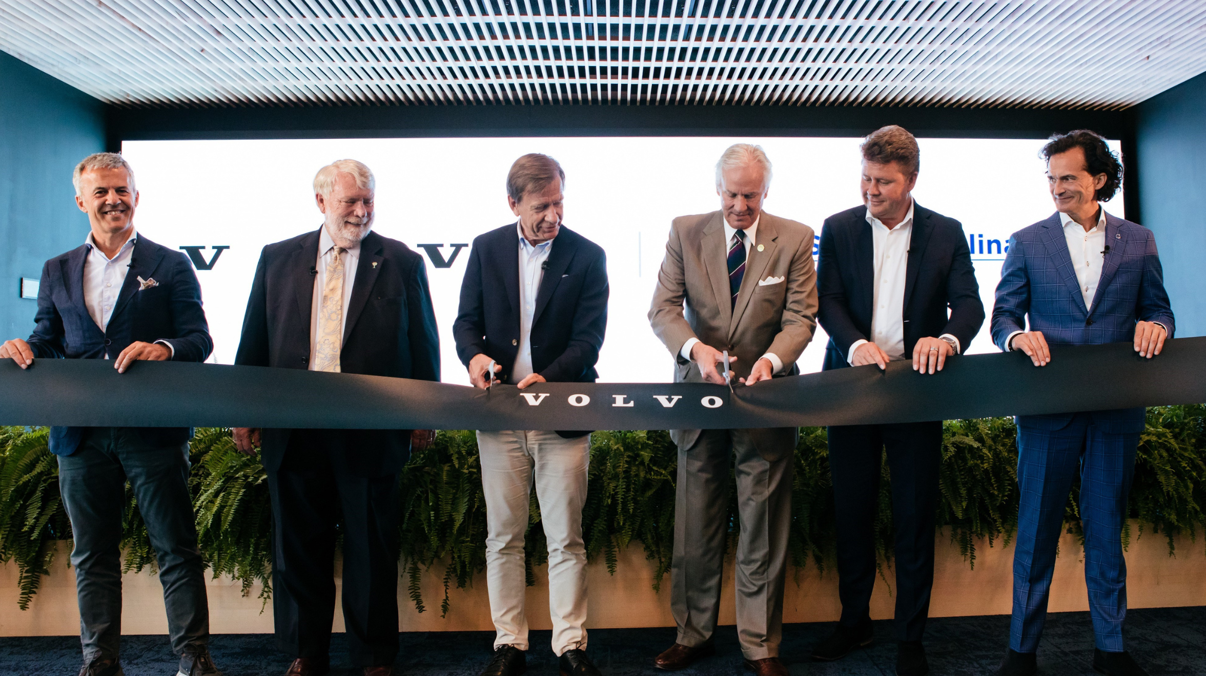 Six people including David Stenström who is the plant manager at Volvo Car Charleston Plant hold up a black ribbon with the word Volvo on it. The two people in the center prepare to cut into the middle of it to celebrate the official opening of Volvo Car University Campus in South Carolina.