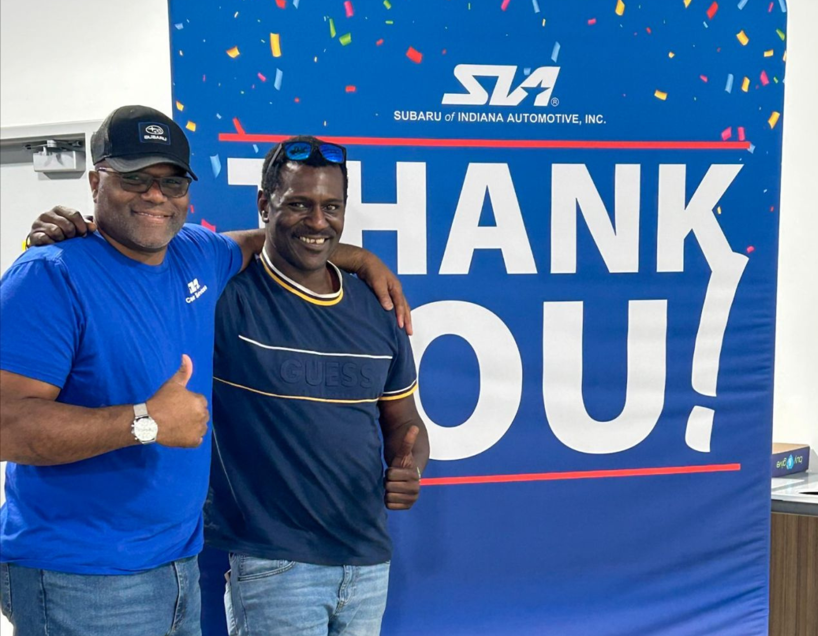 two people giving a thumbs up gesture in front of a Subaru of Indiana Automotive sign that reads thank you