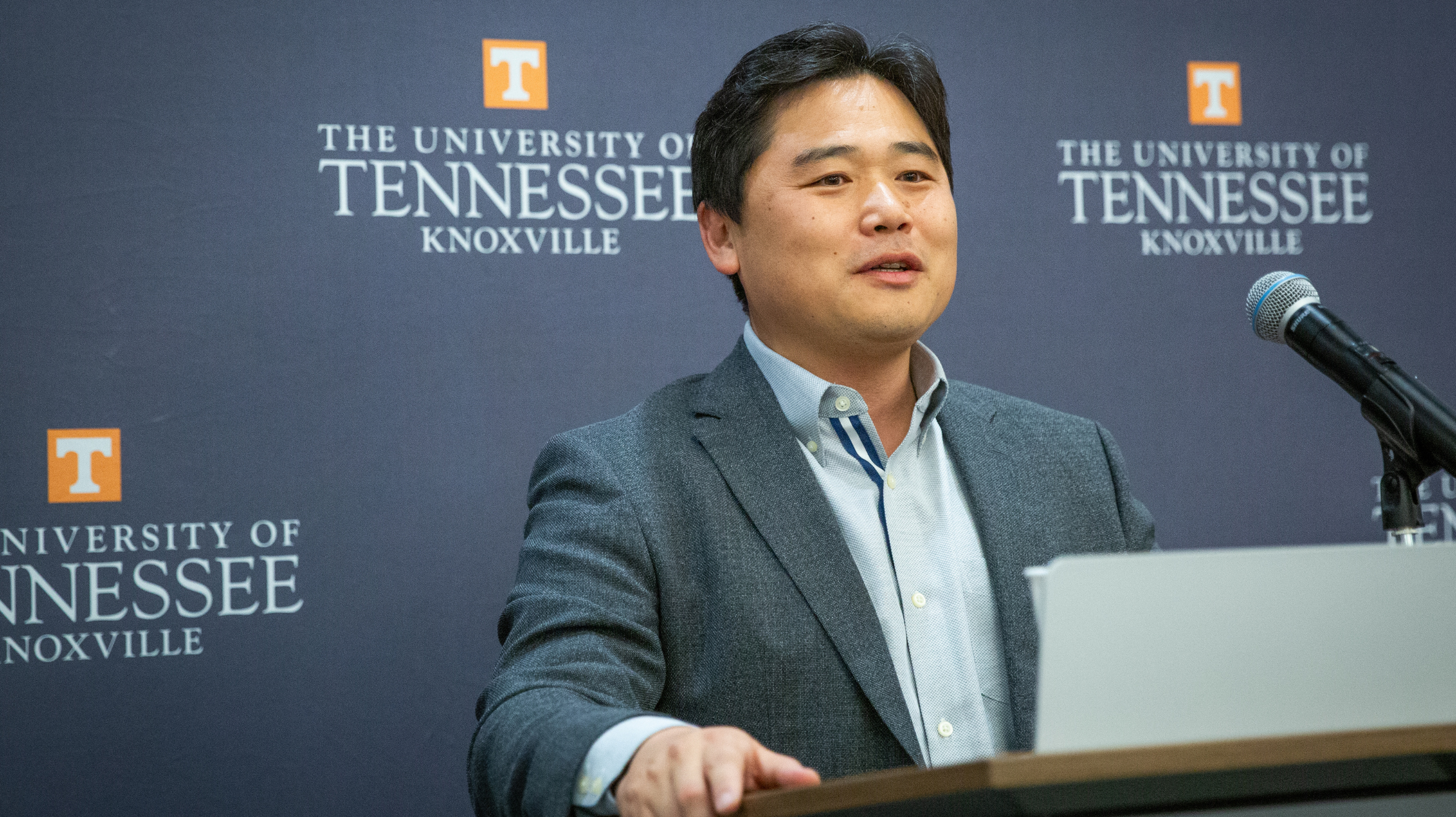 A man stands behind a podium and speaks into a microphone against a backdrop with the University of Tennessee Knoxville logo